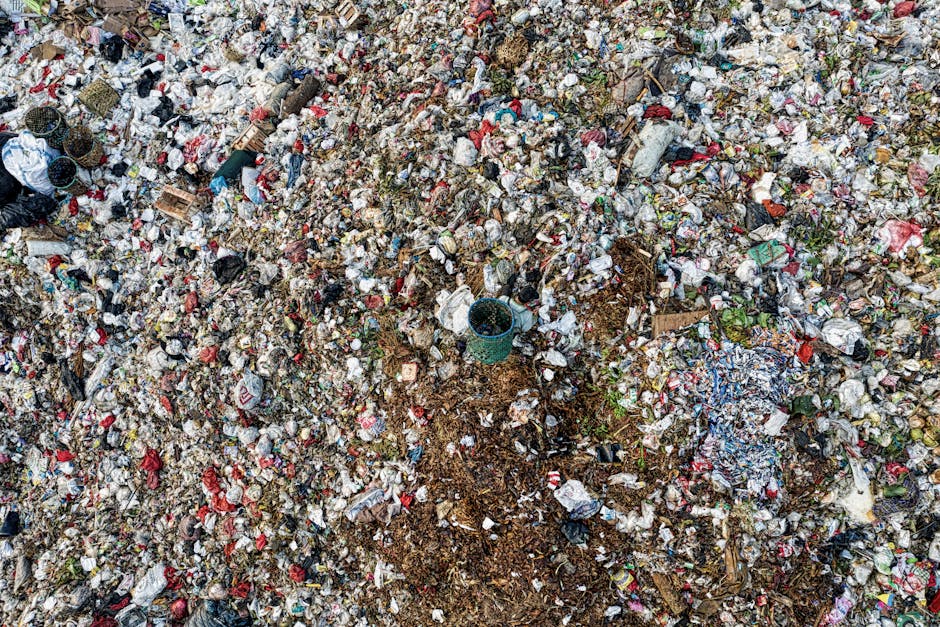 An aerial view of a large, densely packed landfill site filled with mixed waste materials, primarily plastic bottles, paper, cardboard, and food packaging in various colors and textures. The waste app