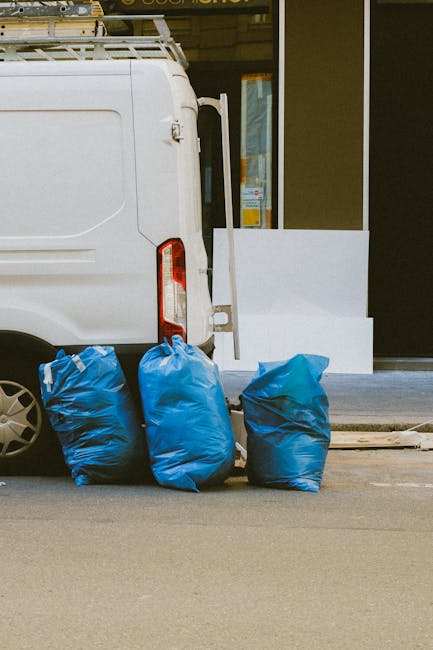 A waste collection operative dressed in green workwear and a helmet stands on the back of a compact rubbish collection vehicle, which is parked on the side of a street lined with trees. The vehicle's 