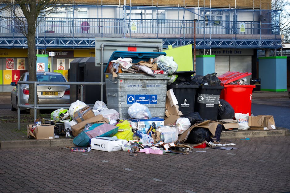 A large green wheeled rubbish bin positioned outdoors on a paved surface against a dark brick wall, filled with red and black plastic bags labeled with garden and outdoor product packaging. The lid of