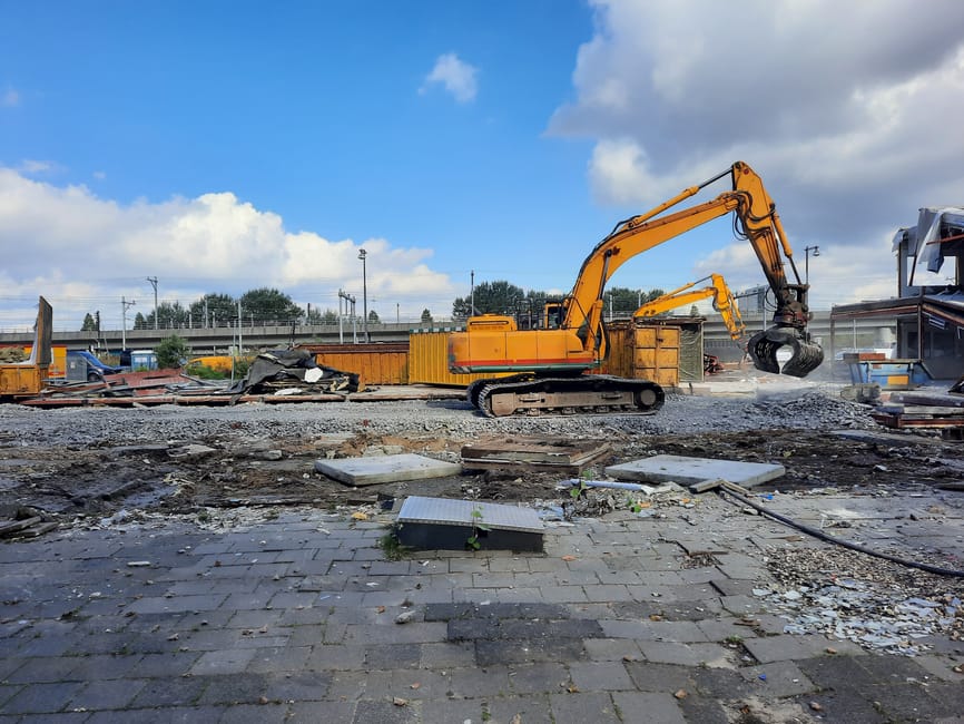The image features an orange excavator equipped with a claw attachment, actively engaged in clearing debris at an outdoor site with a partially dismantled paved surface. The excavator is positioned ce