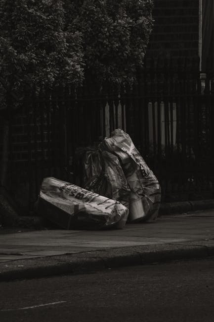 Two large black trash bags filled with waste materials are positioned on the edge of a sidewalk in an outdoor urban setting. The bags appear to be made of thick, crinkled plastic and are partly covere