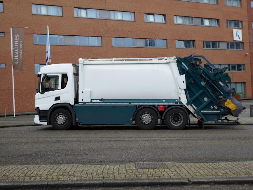 A row of three large black wheelie bins with brightly coloured plastic lids—yellow, blue, and black—placed on a paved sidewalk next to a brick building, with a parked grey van in the background. T